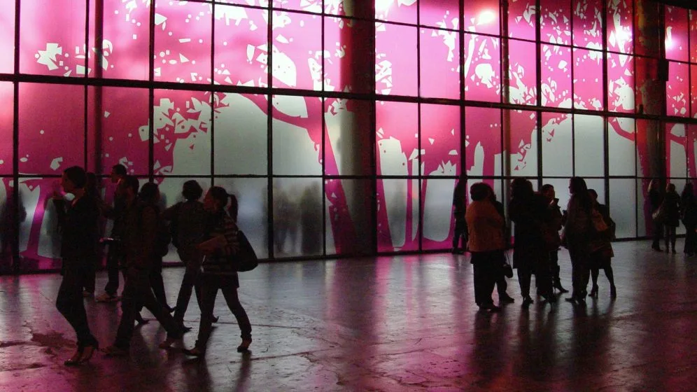 Público circula diante de painel de vidro iluminado em rosa na entrada da São Paulo Fashion Week